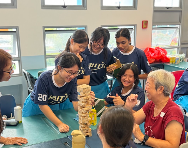 A group of Hai Sing Catholic School students in PE uniforms smiling and interacting with a senior citizen as they carefully play a wooden block tower (Jenga) game together.